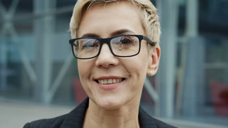 Close-Up-Of-The-Charming-Woman-In-Glasses-With-Short-Hair-Looking-Straight-Into-The-Camera-On-The-Modern-Glass-Building-Background