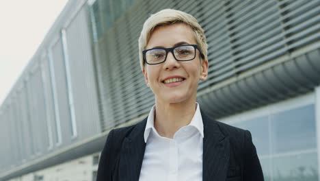 Portrait-Of-The-Succesful-Middle-Aged-Businesswoman-With-Short-Blond-Hair-Turning-Her-Head-To-The-Camera-And-Smiling-On-The-Big-Urban-Building-Background