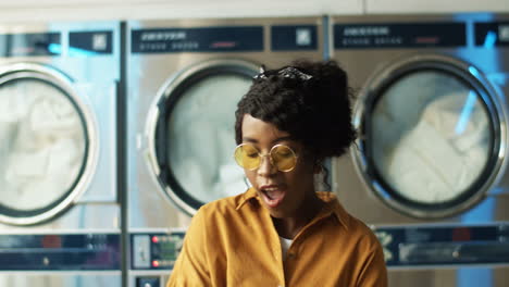 Pretty-Cheerful-Girl-In-Yellow-Glasses-Having-Fun-And-Dancing-In-Laundry-Service-Room-While-Machines-Washing-On-Background