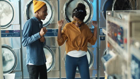 Young-Cheerful-Pretty-Girl-And-Stylish-Handsome-Guy-Having-Fun-And-Dancing-In-Laundry-Service-Room-While-Machines-Washing-On-Background
