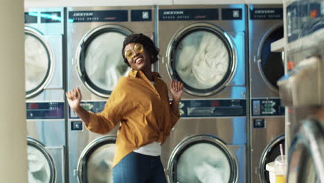 Beautiful-Cheerful-Woman-In-Stylish-Outfit-Having-Fun-And-Dancing-In-Laundry-Service-Room-While-Machines-Washing-On-Background