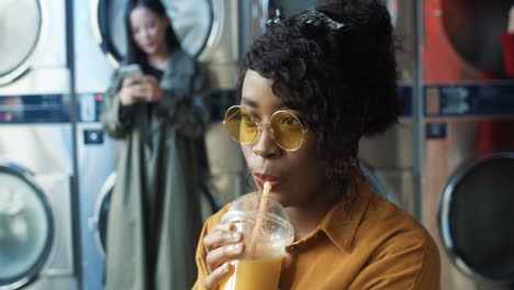 Close-Up-Of-Pretty-And-Happy-Girl-In-Yellow-Glasses-Drinking-Orange-Juice-With-Straw,-Resting-And-Waiting-For-Clothes-To-Be-Washed