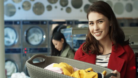 Portrait-Shot-Of-Young-Pretty-Woman-Smiling-To-Camera-And-Holding-Basket-With-Dirty-Clothes-While-Standing-In-Laundry-Service