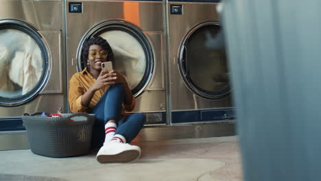 Young-Girl-Sitting-On-Floor-With-Basket-Of-Dirty-Clothes-And-Tapping-On-Smartphone-While-Washing-Machines-Working-Beautiful-Woman-Watching-Video-On-Phone-Or-Chatting-In-Lundry-Service