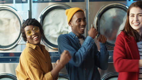 Young-Mixed-Races-Cheerful-Stylish-Girls-And-Guy-Having-Fun-And-Dancing-In-Laundry-Service-Room-While-Machines-Washing-On-Background