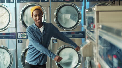 Young-Stylish-Cheerful-Man-Having-Fun-And-Dancing-In-Laundry-Service-Room-While-Machines-Washing-On-Background