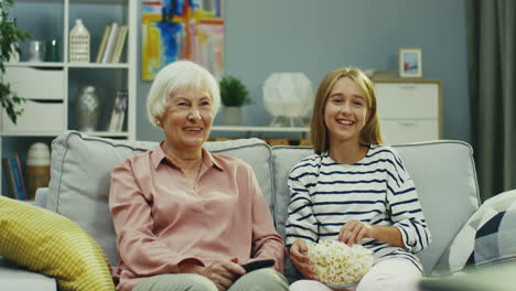 Portrait-Shot-Of-The-Pretty-Small-Teenager-Girl-And-Senior-Grandmother-Sitting-Together-On-The-Sofa-At-Home,-Watching-Tv-And-Laughing