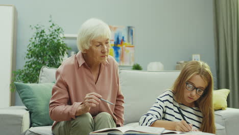 Hübsches-Blondes-Schulmädchen-Mit-Brille,-Das-In-Ein-Schreibheft-Schreibt,-Während-Sie-Ihre-Hausaufgaben-Macht-Und-Ihre-Großmutter-Ihr-Hilft