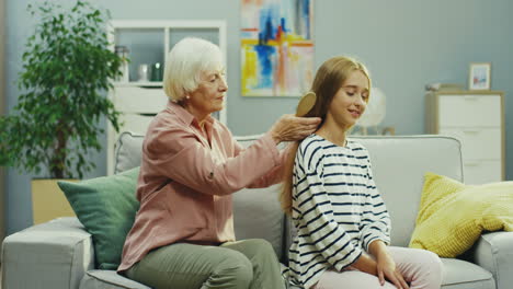 Senior-Woman-Combing-Long-Fair-Hair-Of-Her-Teenager-Granddaughter-On-The-Sofa-In-The-Living-Room