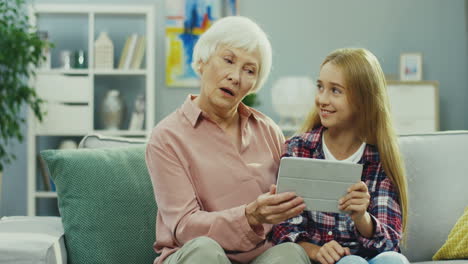 Portrait-Shot-Of-The-Gray-Haired-Grandmother-And-Pretty-Teenager-Granddaughter-Sitting-On-The-Couch-In-The-Cozy-Room-And-Watching-Something-On-The-Screen-Of-The-Tablet-Computer