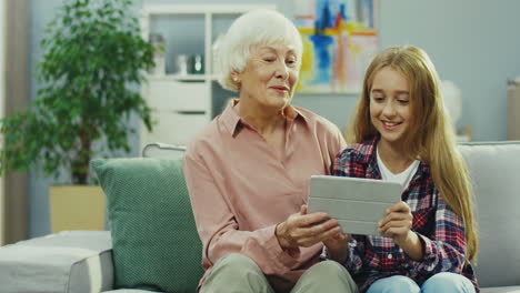 Cheerful-Andsmiled-Grandmother-And-Granddaughter-Sitting-Together-On-The-Sofa-At-Home-And-Watching-Something-On-The-Tablet-Device