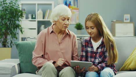 Pretty-Teenager-Girl-Sitting-On-The-Couch-In-The-Nice-Room,-Using-Tablet-Computer-And-Talking-With-Her-Grandmother-Who-Sitting-Next-To-Her