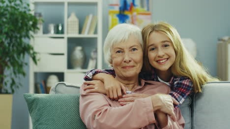 Portrait-Of-The-Happy-Teenager-Girl-Hugging-Her-Grandmother-While-She-Sitting-On-The-Sofa-At-Home-And-They-Smiling-To-The-Camera