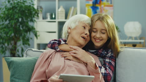 Grauhaarige-Seniorin,-Die-Mit-Einem-Tablet-Gerät-Auf-Der-Couch-Sitzt,-Und-Ihre-Süße-Teenager-Enkelin-Kommt-Von-Hinten-Und-Umarmt-Sie