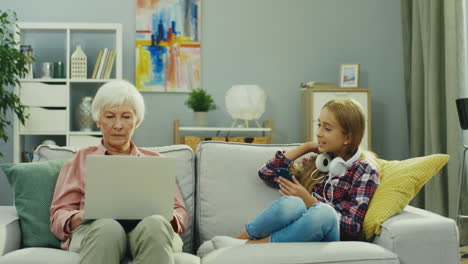 Old-Woman-Sitting-On-The-Sofa-And-Working-On-The-Laptop-Computer-While-Her-Granddaughter-Sitting-Beside-And-Listening-To-The-Music-,Then-Girl-Taking-Off-Them-And-Talking-With-Grandma