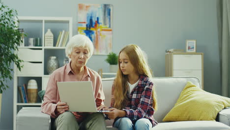 Old-Good-Looking-Woman-Sitting-On-The-Couch-In-The-Cozy-Livingroom-And-Telling-Something-To-Her-Pretty-Teen-Granddaughter-While-Demonstrating-Something-On-The-Laptop-Screen