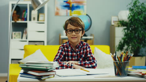 Portrait-Shot-Of-The-Cute-Small-Boy-In-Glasses-Sitting-At-The-Desk-With-School-Stuff-And-Textbooks-In-His-Room-And-Smiling-To-The-Camera