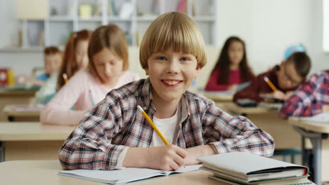 Close-Up-Of-The-Cute-Small-Teen-Schoolboy-In-The-Classroom-Writing-With-A-Pencil-In-The-Copybook,-Then-Smiling-Cheerfully-To-The-Camera