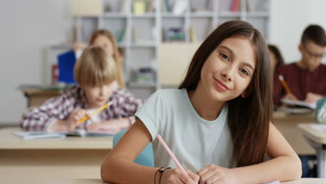 Portrait-Shot-Of-The-Little-Teen-Girl-Writing-In-The-Copybook-During-Test-At-The-Lesson-In-The-Classroom-And-Smiling-To-The-Camera