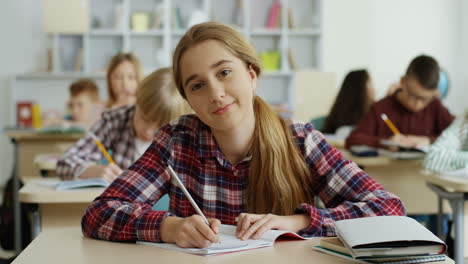 Portrait-Of-The-Teen-Pretty-Schoolgirl-With-Fair-Hair-Writing-In-The-Copybook-At-The-Lesson-In-The-Classroom-And-Smiling-To-The-Camera