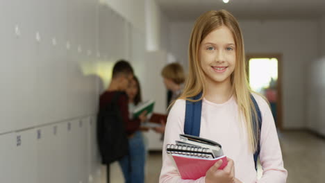 Portrait-Shot-Of-The-Teenage-Pretty-Schoolgirl-With-Textbooks-And-Schoolbag-Standing-At-The-School-Corridor-And-Smiling-To-The-Camera