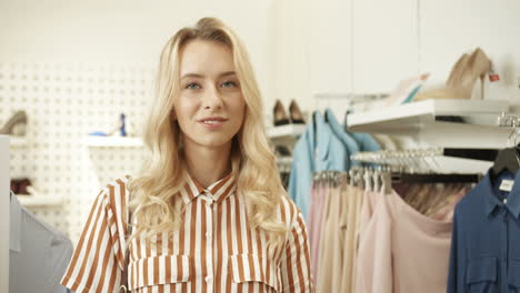 Portrait-Of-Young-Blonde-Happy-Woman-With-Curly-Hair-Smiling-Cheerfully-To-Camera-In-Clothing-Shop