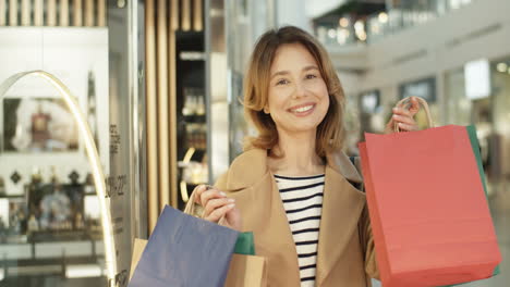 Portrait-Of-Charming-Pretty-Woman-In-Coat-Standing-At-Shopping-Mall-Passage-And-Smiling-Cheerfully-While-Demonstrating-Bags