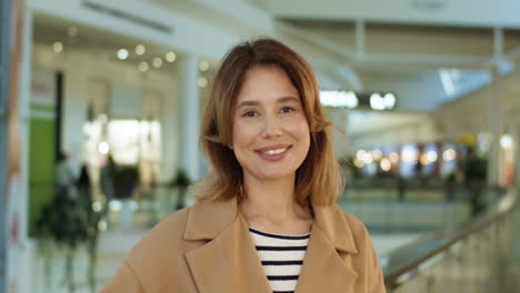 Portrait-Of-Attractive-Woman-In-Coat-Standing-At-Shopping-Mall-Passage,-Turning-Face-And-Smiling-Cheerfully-To-Camera