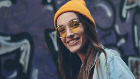 Back-View-Of-The-Close-Up-Of-The-Hipster-Stylish-Young-Girl-In-Hat-And-Sunglasses-Turning-Her-Head-Smiling-Cheerfully-To-The-Camera-On-The-Graffity-Background