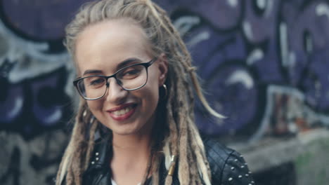 Close-Up-Of-The-Camera-Zooming-In-The-Face-Of-The-Pretty-Stylish-Girl-In-Glasses-And-With-Dreadlocks-Standing-On-The-Graffity-Wall-Background-And-Smiling