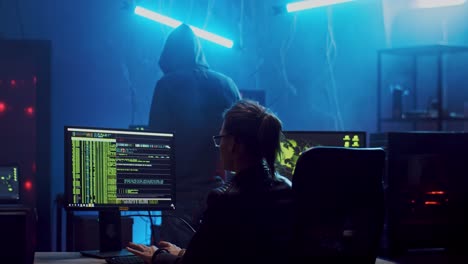 Rückseite-Der-Jungen-Hackerin-Mit-Brille,-Die-Nachts-Am-Computer-Arbeitet-Und-Ein-Programm-Hackt,-Während-Ihr-Männlicher-Kollege-In-Einer-Kapuze-Durch-Den-Raum-Geht