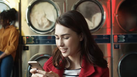 Pretty-Stylish-Woman-Tapping-And-Texting-Message-On-Smarphone-While-Sitting-In-Laundry-Service-Room
