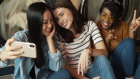 Drei-Hübsche-Gemischte-Rassen-Freundliche-Mädchen-An-Waschmaschinen-Im-Waschhaus-Lächeln-Zur-Smartphone-kamera-Und-Machen-Selfire-foto