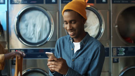 Stylish-Man-In-Yellow-Hat-Tapping-And-Texting-Message-On-Smarphone-While-Sitting-In-Laundry-Service-Room