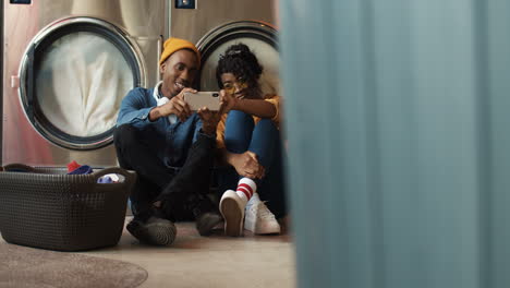 Young-Couple-Sitting-On-Floor-With-Basket-Of-Dirty-Clothes-And-Watching-Video-On-Smartphone-While-Washing-Machines-Working-Man-And-Woman-Using-Phone-And-Resting-In-Lundry-Service