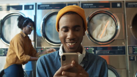 Close-Up-Of-Stylish-Man-In-Yellow-Hat-Tapping-And-Texting-Message-On-Smarphone-While-Sitting-In-Laundry-Service-Room
