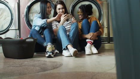 Mixed-Races-Girls-Friends-Sitting-On-Floor-With-Basket-Of-Dirty-Clothes-And-Watching-Video-On-Smartphone-While-Washing-Machines-Working