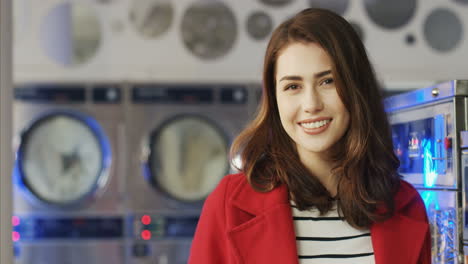 Close-Up-Of-Beautiful-Young-Woman-In-Red-Coat-Smiling-Cheerfully-To-Camera-In-Laundry-Service-Room