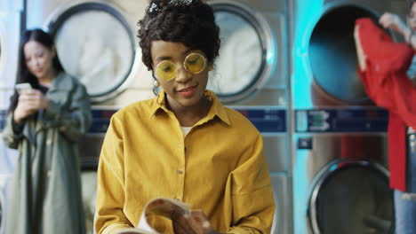 Young-Pretty-And-Stylish-Girl-In-Yellow-Glasses-Standing-In-Laundry-Service-Room-And-Flipping-Pages-Of-Fashion-Journal
