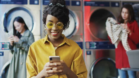 Young-Pretty-And-Stylish-Girl-In-Yellow-Glasses-Standing-In-Laundry-Service-Room-And-Tapping-On-Smartphone