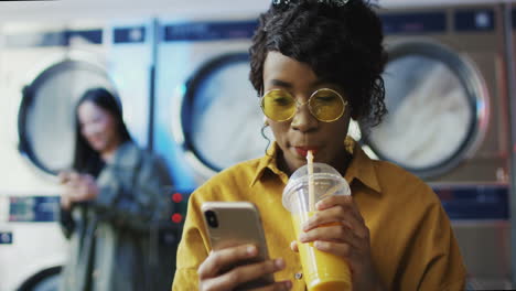 Girl-Drinking-Orange-Juice-With-Straw,-Texting-Message-On-Phone-And-Waiting-For-Clothes-To-Get-Clean-Woman-Sipping-Drink-In-Laundry-Service-Room-And-Tapping-Or-Scrolling-On-Smartphone