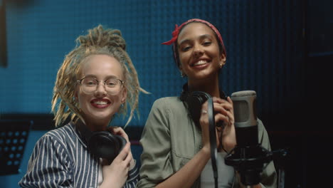 Close-Up-Of-The-Two-Charming-And-Cheerful-Young-Female-Singers-Finishing-Song-Recording-At-The-Studio,-Taking-Off-Headphones-And-Smiling-To-The-Camera