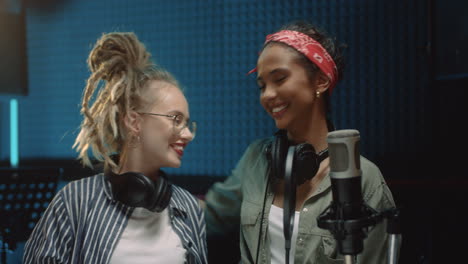 Portrait-Of-The-Two-Happy-Beautiful-And-Stylish-Young-Women-Singers-Standing-In-The-Sound-Studio-Room-At-The-Mic,-Hugging-And-Smiling-Cheerfully-To-The-Camera
