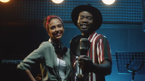 Portrait-Of-The-Multiethnic-Stylish-Young-Man-And-Woman,-Singers-Of-The-Duo-Looking-At-Each-Other-And-Then-Smiling-Cheerfully-To-The-Camera-In-The-Dark-Sound-Studio-During-Recording