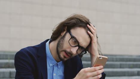 Portrait-Of-The-Young-Sad-Businessman-In-Glasses-Looking-Worried-While-Texting-And-Chatting-On-The-Smartphone