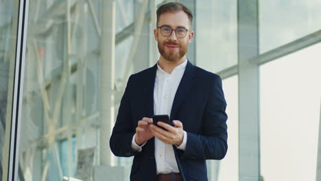 Portrait-Shot-Of-The-Young-Handsome-Businessman-Taping-And-Texting-On-The-Smartphone-In-His-Hands,-Then-Rising-Head-And-Smiling-To-The-Camera