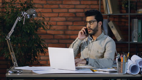Good-Looking-Young-Man-Architect-Or-Designer-In-Glasses-Talking-On-The-Phone-While-Sitting-At-The-Laptop-Computer-In-The-Office-Room