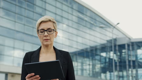 Portrait-Of-The-Businesswoman-In-Glasses-And-With-Short-Hair-Taping-On-The-Tablet-Computer-Near-Big-Modern-Office-Building