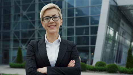 Cheerful-Woman-In-The-Glasses-And-With-Short-Blond-Hair-Looking-At-The-Side-And-Then-Turning-Her-Head-To-The-Camera-With-A-Smile-On-The-Big-Glass-Building-Background