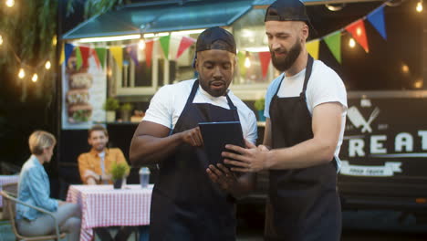 Two-Cheerful-Male-Waiters-In-Aprons-And-Caps-Standing-Outdoor-Foodd-Truck-At-Park-Festival